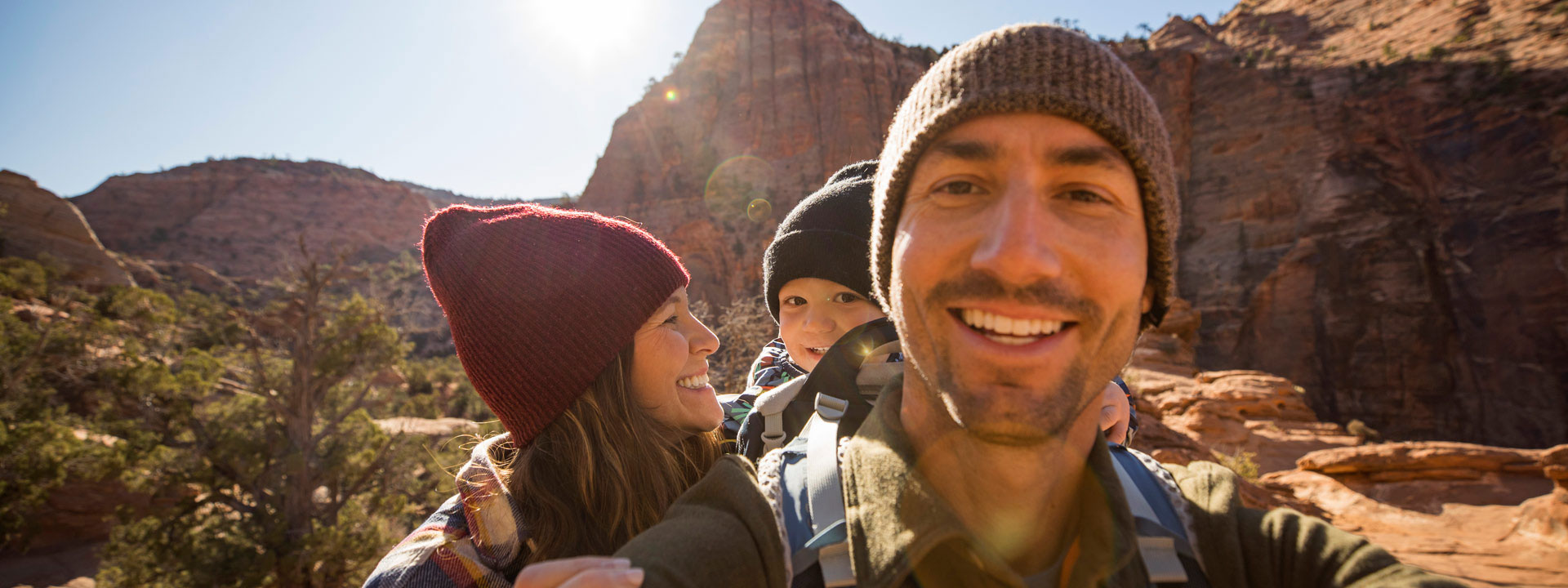 A smiling family of three wearing beanies, hiking through a sunlit canyon with red rock formations, conveying joy and adventure in the outdoors.