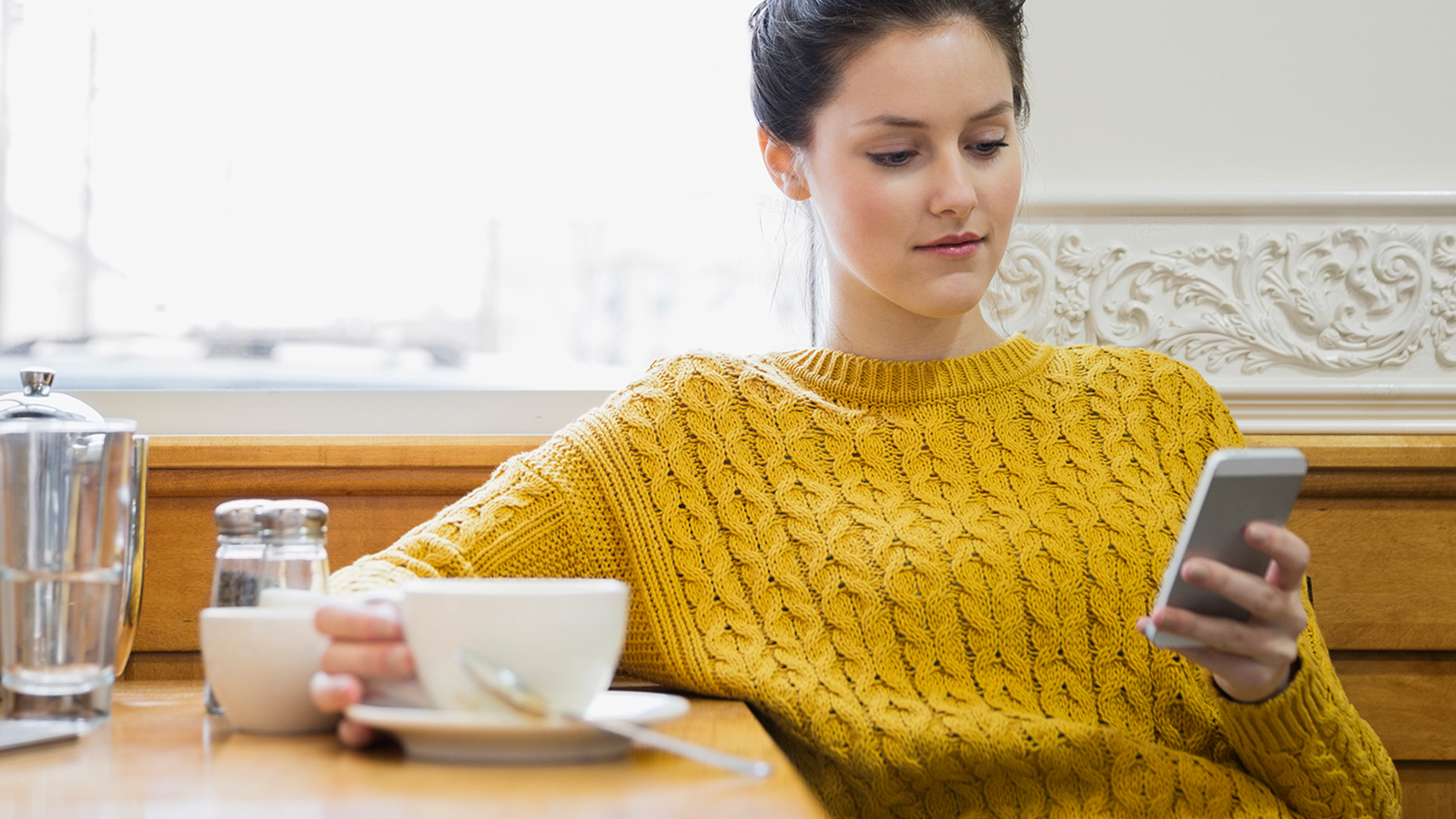 A woman sitting in a cafe holding a cup in right hand and viewing a mobile phone held in her left hand.