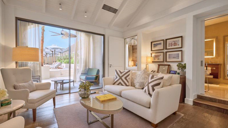 Cozy living room with a white couch, patterned cushions, and an armchair. A sliding glass door opens to a sunny patio. Warm lighting adds a welcoming feel.