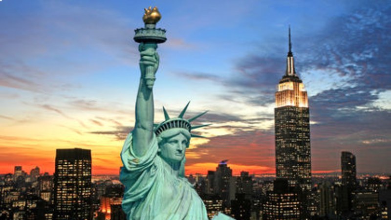 Statue of Liberty with New York City skyline and Empire State Building at sunset.