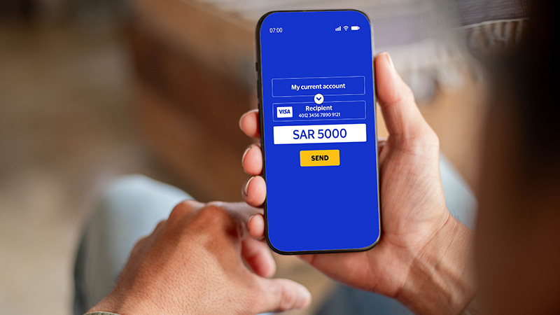 A person holds a smartphone displaying a banking app interface. The screen shows a payment of SAR 5000 to a Visa recipient, with a "Send" button below.