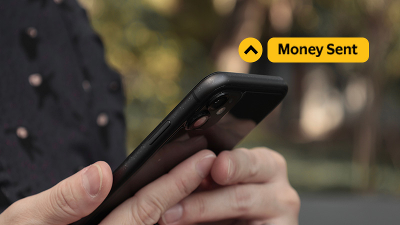 A person holds a smartphone outdoors, focusing on the screen. A yellow notification displays "Money Sent," conveying a successful transaction.